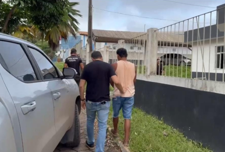 Imagem do suspeito sendo coduzido por dois agentes até a delegacia após a prisão, ao lado de uma caminhonete cinza.