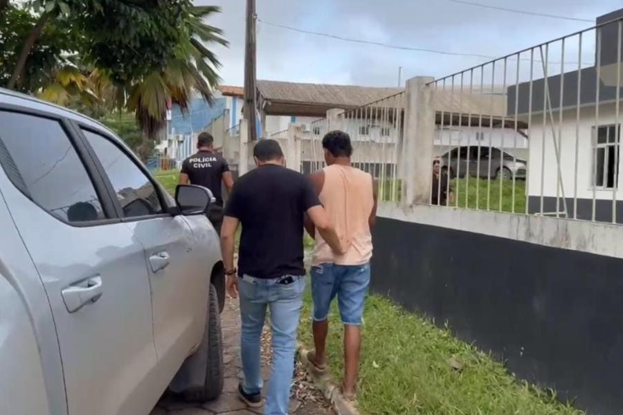 Imagem do suspeito sendo coduzido por dois agentes até a delegacia após a prisão, ao lado de uma caminhonete cinza.